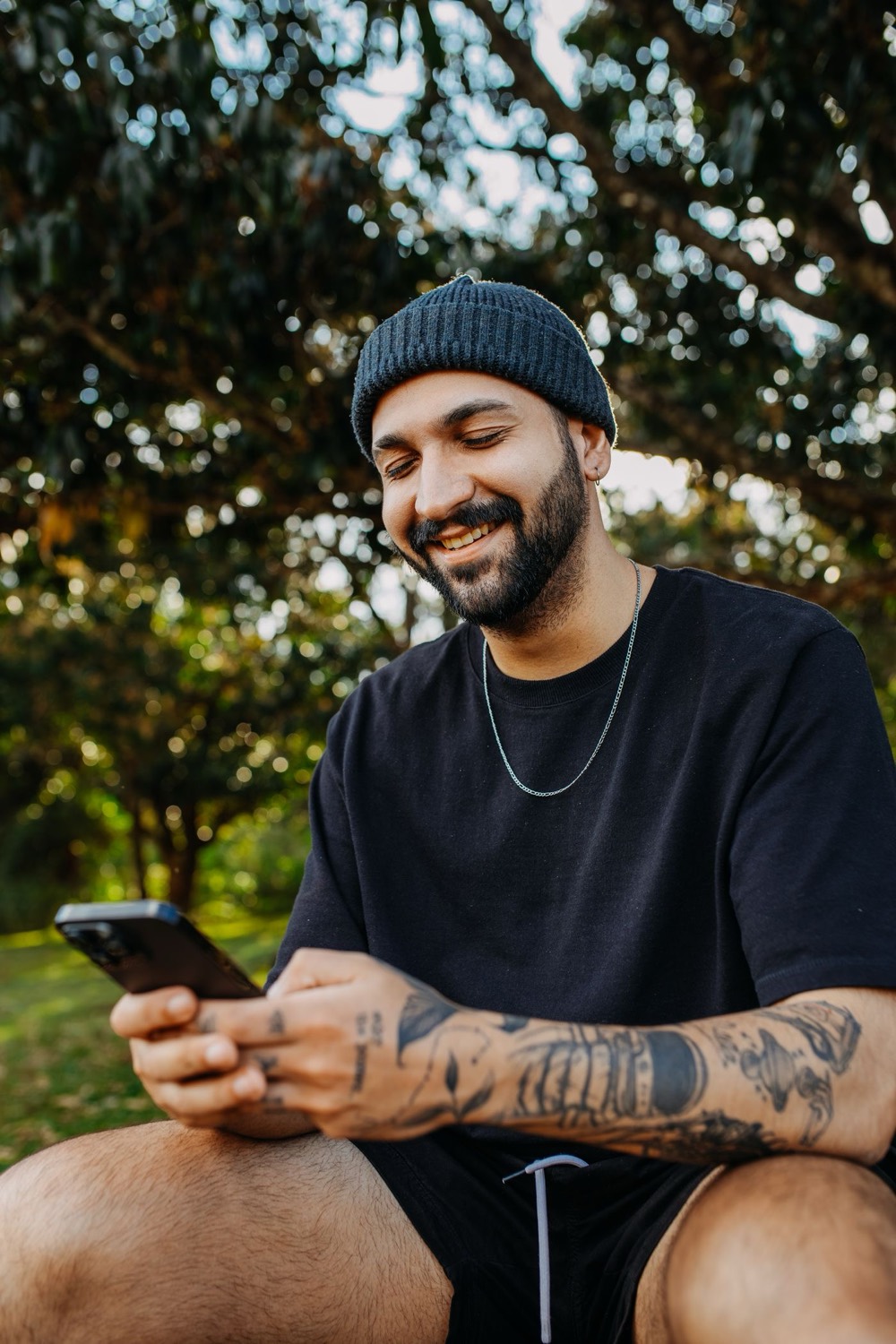 A smiling young man in a beanie sitting outdoors under trees, checking a payment on his phone